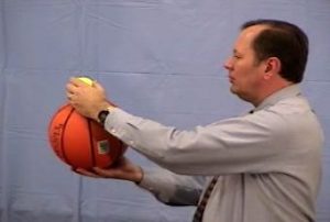 The instructor holds a tennis ball on top of a basketball at chest level.